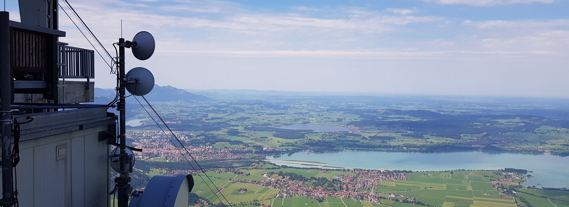 Das Foto zeigt das Voralpenland von der Bergstation der Tegelbergbahn aus. Man sieht unterschiedlichen Ortschaften, die um einen großen See angesiedelt sind. Man blickt in die weite Ebene, die mit Wäldern, Wiesen und weiteren vereinzelten kleinen Ortschaften durchzogen ist.  Im Vordergrund sieht man einen Teil der Bergstation  und die Tragseile der Bergbahn die in die Tiefe führen. 