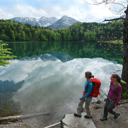 Das Bild zeigt zwei Frauen, die Wanderkleidung und Rucksäcke tragen. Sie stehen am Ufer eines Sees. Die Wasseroberfläche des Sees ist ganz ruhig. Der See ist mit hohen Bäumen eingerahmt. Im Hintergrund erheben sich die Berge. Der Himmel ist blau. Wolken ziehen auf.   