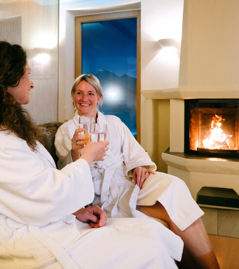 Das Bild zeigt zwei Frauen, die auf einem Sofa vor einem Kamin sitzen. Im Kamin brennt ein Feuer hinter einer Glasscheibe. Die Frauen tragen weiße Bademäntel und prosten sich mit Wassergläsern zu. Sie lächeln sich an. 