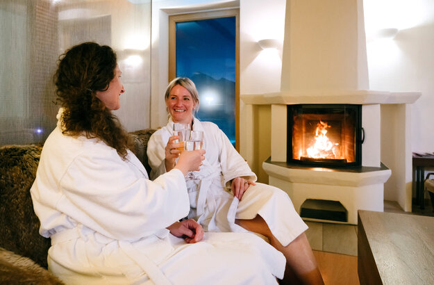 Das Bild zeigt zwei Frauen, die auf einem Sofa vor einem Kamin sitzen. Im Kamin brennt ein Feuer hinter einer Glasscheibe. Die Frauen tragen weiße Bademäntel und prosten sich mit Wassergläsern zu. Sie lächeln sich an. 