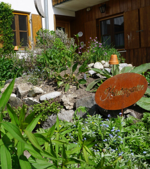 Das Bild zeigt einen wild wuchernden Kräutergarten, in dem es wächst, blüht und gedeiht. Zwischen den Pflanzen steckt ein Metallschild, auf dem Kräutergarten zu lesen ist. Im Hintergrund ist die Wand eines Hauses mit hölzernen Fensterläden zu erkennen.