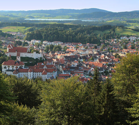 Das Bild zeigt Füssen von oben. Es ist von einer Anhöhe aus aufgenommen. Über grüne Baumwipfel hinweg erblickt man die roten Dächer der Stadt. Im Vordergrund sind die großen Gebäudekomplexe des ehemaligen Benediktinerklosters St. Mang und des Hohen Schlosses zu sehen. Im Hintergrund erheben sich bewaldete Hügel. Davor liegt ein blauer See.