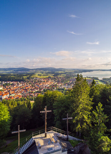 Das Bild zeigt einen Berg mit drei Kreuzen, die auf einer steinernen Aussichtsplattform stehen, aus der Vogelperspektive. Die Aussichtsplattform ist umgeben von hohen, grünen Bäumen. Am Fuß des Berges ist eine Stadt und Seen zu sehen.