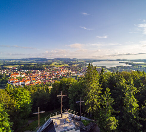Das Bild zeigt einen Berg mit drei Kreuzen, die auf einer steinernen Aussichtsplattform stehen, aus der Vogelperspektive. Die Aussichtsplattform ist umgeben von hohen, grünen Bäumen. Am Fuß des Berges ist eine Stadt und Seen zu sehen.