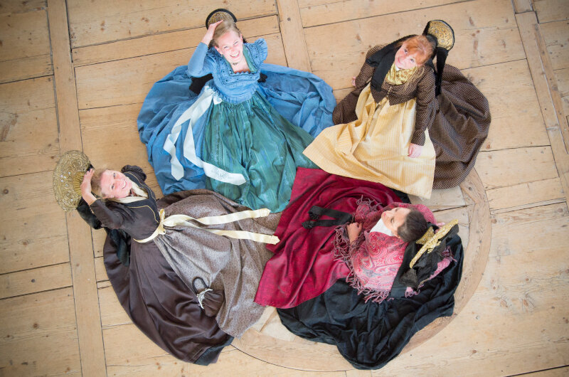 Fünf Frauen, gekleidet in historische, farbenfrohe Kleider, sitzen im Kreis auf einem Holzfußboden. Jede trägt einen auffälligen Hut und lächelt. Die Kleider sind in verschiedenen Farben, darunter Blau, Braun, Rot und Gelb, und sie posieren gemeinsam für die Aufnahme.