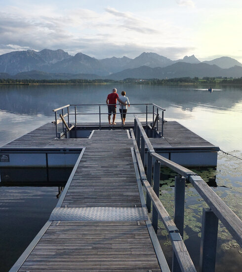 Das Bild zeigt einen großen Steg, der in den Hopfensee ragt. Auf dem See schwimmen Seerosenblätter. Am Ende des Stegs wird öffnet sich eine Plattform, in deren Mitte ein Kneipptretbecken eingelassen ist. Auf der Kneippinsel stehen zwei Menschen mit dem Rücken an ein Geländer gelehnt und blicken in die Ferne. Der Himmel ist in ein ruhiges Abendlicht getaucht und im Hintergrund erhebt sich die Bergkette der Allgäuer Alpen.