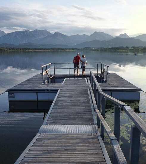 Das Bild zeigt einen großen Steg, der in den Hopfensee ragt. Auf dem See schwimmen Seerosenblätter. Am Ende des Stegs wird öffnet sich eine Plattform, in deren Mitte ein Kneipptretbecken eingelassen ist. Auf der Kneippinsel stehen zwei Menschen mit dem Rücken an ein Geländer gelehnt und blicken in die Ferne. Der Himmel ist in ein ruhiges Abendlicht getaucht und im Hintergrund erhebt sich die Bergkette der Allgäuer Alpen.