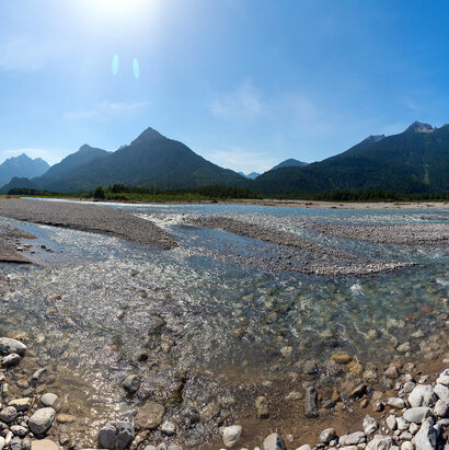 Auf dem Bild sieht man ein Flussbett umrahmt von Wald. Im Hintergrund sind Berge zu erkennen. Sie Sonne strahlt über das Bild.
