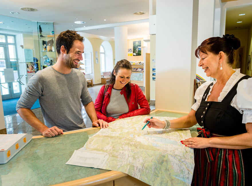 Das Bild zeigt einen Mann und zwei Frauen, die an einem Informationstresen stehen. Der Mann und die Frau, die hinter dem Tresen stehen, tragen Freizeitkleidung. Die Dame auf der anderen Seite trägt ein Dirndl mit einer grün-roten Schürze. Alle lächeln. Die Frau im Dirndl deutet mit einem Stift auf eine Karte, die vor den Personen auf dem Tresen liegt. Alle lächeln.