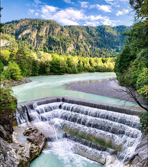 Das Bild zeigt einen atemberaubenden Blick auf einen Wasserfall – den Lechfall bei Füssen, der sich über eine felsige Klippe hinunterstürzt. Der Wasserfall, mit seinem weißen Schaum, ist der Hauptakzent im Bild. Er ist von üppig grünen Bäumen und Büschen umgeben, die der Szene eine Prise der Naturpracht verleihen. Der Wasserfall liegt in einem Tal, durch das der Fluss Lech fließt. Der klare blaue Himmel über ihm ist von fluffigen, weißen Wolken durchzogen. Die Perspektive des Bildes ist aus einer hohen Position, was eine Panoramaansicht des Wasserfalls und seiner Umgebung bietet.