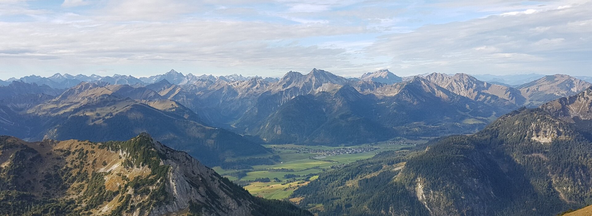 Man blickt auf unzählige Berggipfel, die sich vor dem Betrachter erheben. Dazwischen ist ein grünes Tal zu sehen. Dort liegt eine kleine Ortschaft. Der Himmel ist blau, jedoch von weißen Wolken durchzogen.  
