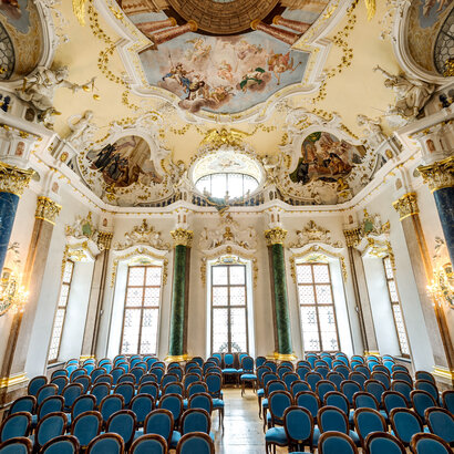 Das Foto zeigt eine prunkvollen Raum. Die Decke ist reich mit Stuck und mehreren Deckengemälden verziert. Den Raum säumen hohe Fenster. Auch die Fensterrahmen sind mit weiß, goldenem Stuck verziert. Der Raum wird von vier Säulen eingerahmt. Zwei Säulen sind Lapislazuli blau, zwei malachitgrün. Der Raum steht voller Holzstühle, die mit blauem Stoff bezogen sind. Die Stühle sind alle Richtung Kamera ausgerichtet. Ein schmaler Gang führt durch die Stühle hindurch. Der Raum ist lichtdurchflutet.  