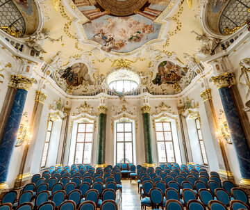 Das Foto zeigt eine prunkvollen Raum. Die Decke ist reich mit Stuck und mehreren Deckengemälden verziert. Den Raum säumen hohe Fenster. Auch die Fensterrahmen sind mit weiß, goldenem Stuck verziert. Der Raum wird von vier Säulen eingerahmt. Zwei Säulen sind Lapislazuli blau, zwei malachitgrün. Der Raum steht voller Holzstühle, die mit blauem Stoff bezogen sind. Die Stühle sind alle Richtung Kamera ausgerichtet. Ein schmaler Gang führt durch die Stühle hindurch. Der Raum ist lichtdurchflutet.