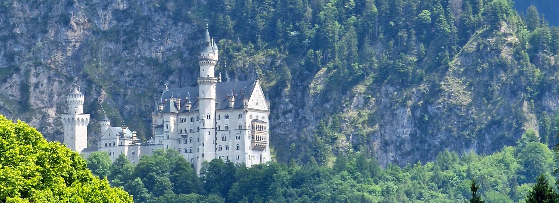 Das Bild zeigt eine Frühlingsszene. Auf einer verschneiten, bewaldeten Anhöhe steht das weiße Schloss Neuschwanstein. Hinter dem Schloss erhebt sich teilweise bewaldeter Berg. Zwischen den Bäumen ist auch immer wieder der rohe Fels zu sehen. Die Sonne scheint. Der Himmel ist wolkenverhangen. 
