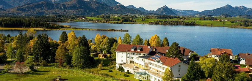 Das Bild zeigt ein großes weißes Haus, das umsäumt von Bäumen am Ufer eines Sees steht. Im Vordergrund sind grüne Felder und im Hintergrund die Gipfel der Allgäuer Alpen zu sehen.
