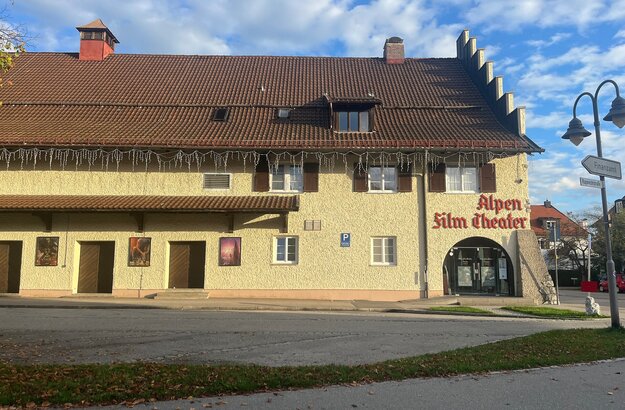 Das Bild zeigt ein Gebäude mit einer beigefarbenen Fassade und einem braunen Dach. Über der Eingangstüre steht in roter Schrift Alpenfilmtheater.