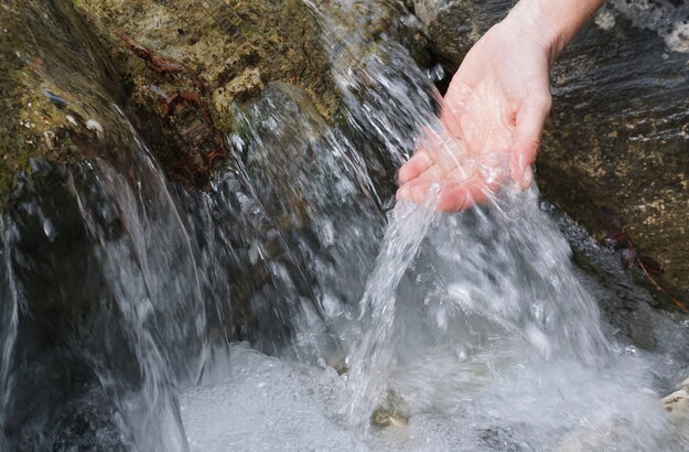 Das Bild zeigt eine Nahaufnahme der Hand einer Person, die unter einem Wasserlauf gehalten wird. Das Wasser aus dem Bach fließt über einen felsigen Absatz in einem kleinen Wasserfall sprudelnd nach unten. 