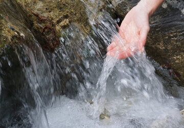 Das Bild zeigt eine Nahaufnahme der Hand einer Person, die unter einem Wasserlauf gehalten wird. Das Wasser aus dem Bach fließt über einen felsigen Absatz in einem kleinen Wasserfall sprudelnd nach unten. 