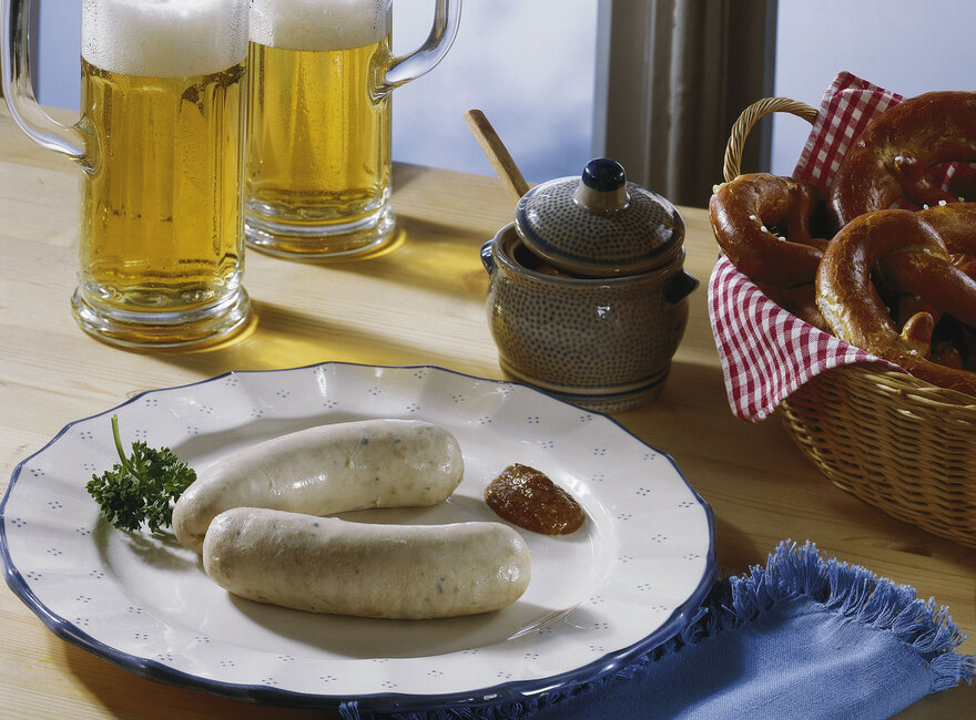 Ein Teller mit Weißwürsten und süßem Senf steht auf einem Tisch. Im Hintergrund befinden sich Bierkrüge und ein Korb mit Brezeln.