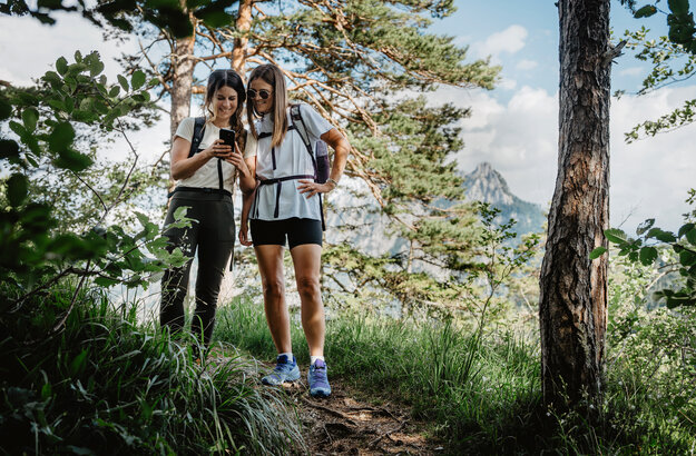 Das Bild zeigt zwei junge Frauen in leichter sportlicher Kleidung und Rucksäcken in einem lichten Kiefernwaldstück. Sie blicken interessiert auf ein Smartphone in ihren Händen.