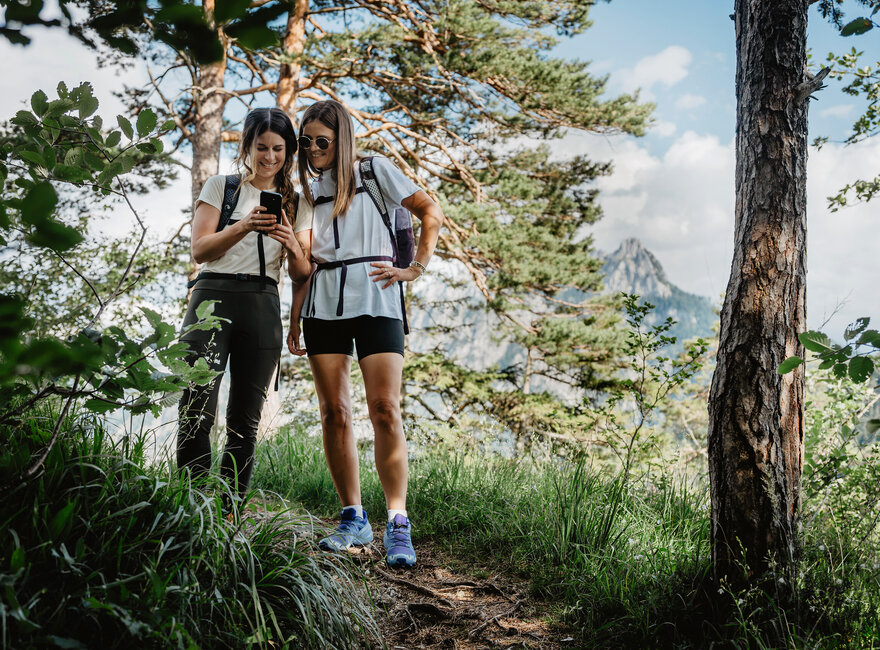 Das Bild zeigt zwei junge Frauen in leichter sportlicher Kleidung und Rucksäcken in einem lichten Kiefernwaldstück. Sie blicken interessiert auf ein Smartphone in ihren Händen.