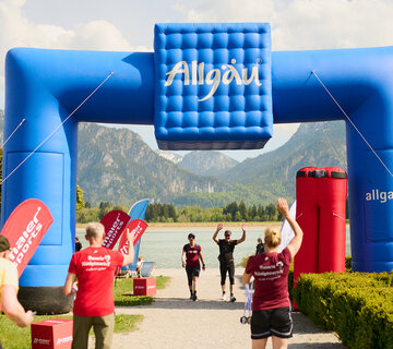 Das Bild zeigt einen großen Torbogen mit dem blauen Allgäu-Würfel vor einer traumhaften Berg- und Seenkulisse, in die sich das Märchenschloss Neuschwanstein einschmiegt. Menschen in Wanderbekleidung stehen vor und laufen durch den Bogen.