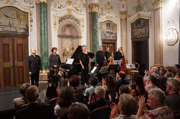 Das Bild zeigt eine Gruppe Musiker auf einer niedrigen Bühne stehen. Einige haben Geigen in den Händen. Auch ein Cello und ein Kontrabass ist dabei. Im Hintergrund steht ein Konzertflügel.  Die Wände des Raums sind prunkvoll verziert. Vor der Bühne sitzen zahlreiche Personen auf Stühlen der Bühne zugewandt und applaudieren. Der Raum ist hell erleuchtet. 