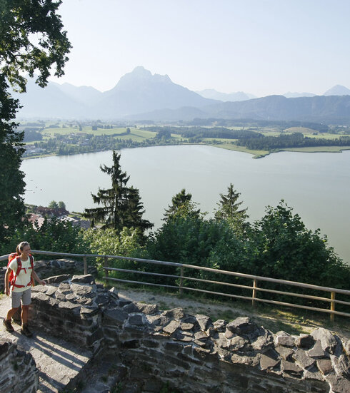 Das Bild zeigt eine friedvolle Szene von zwei Menschen, einem Mann und einer Frau, die in den alten Mauern einer verfallenen Burg herumgehen. Die Ruine befindet sich auf einer Anhöhe über dem Hopfensee und ist von einer lichten Berglandschaft und einem dichten Wald umgeben. Die Bergkette, majestätisch und hoch, bildet eine beeindruckende Kulisse für diese Szenerie. Der Himmel ist ein helles Blau, was zu der allgemeinen Ruhe des Bildes beiträgt. Die beiden Menschen, in legerer Kleidung, scheinen einen gemütlichen Spaziergang am Ufer zu genießen. Das Bild vermittelt ein Gefühl von Frieden und Ruhe und ist somit ein faszinierender Anblick.