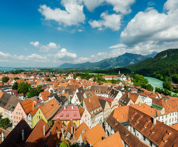 Das Bild zeigt eine Luftaufnahme der Stadt Füssen. Man sieht eine Vielzahl von sich aneinanderreihenden Dächern der Altstadt. Die Farben der größtenteils spitzen Giebeldächer reichen von hellem rot bis hin zu dunklem grau. Die Fassaden der Häuser sind größtenteils weiß, aber auch bunte Fassaden in Blau-, Gelb- oder Grüntönen sind zu sehen. An der Stadt entlang schlängelt sich das türkise Wasser des Flusses Lech. Im Hintergrund erheben sich majestätische Berge gegen den Himmel, deren Spitzen scheinbar die Wolken berühren. Der Himmel über ist Stadt ist blau mit der ein oder andern flauschig weißen Wolke. Das Wetter ist sonnig.