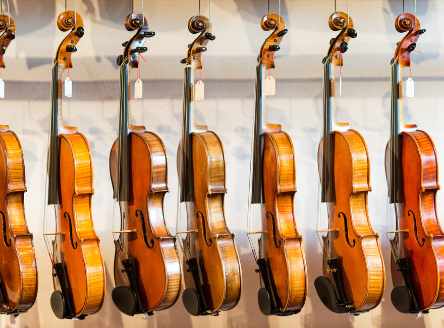 Das Bild zeigt eine Sammlung von Geigen, die jeder einzeln von einem Haken an einer Wand hängt. Die Geigen sind in einer Reihe angeordnet, mit jedem Instrument gleich in eine Richtung zeigend. Die Geigen sind unterschiedlicher Größe, was darauf hindeutet, dass sie für verschiedene Altersgruppen oder Spielweisen konzipiert sind. Die Geigen sind aus Holz und haben eine reiche, warme Braunfärbung, die vor dem weißen Hintergrund im Bild gut zur Geltung kommt. Die Saiten der Geigen sind sichtbar und tragen dazu bei, die Gesamtästhetik des Bildes zu vervollständigen. Die Anordnung der Geigen und deren Positionierung an der Wand erzeugen ein Gefühl von Ordnung und Harmonie