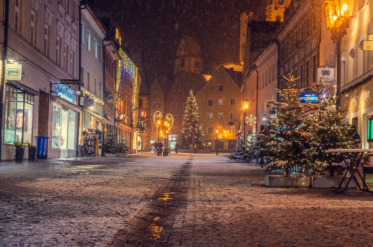 Schneefall bei Nacht in der gepflasterten Reichenstraße in der Füssener Altstadt. Weihnachtsbäume und geschmückte Fassaden.