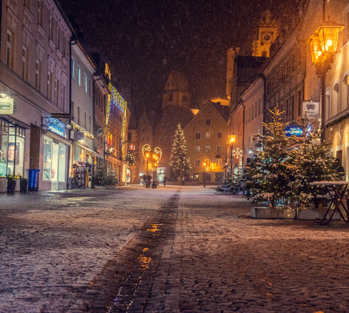 Schneefall bei Nacht in der gepflasterten Reichenstraße in der Füssener Altstadt. Weihnachtsbäume und geschmückte Fassaden.