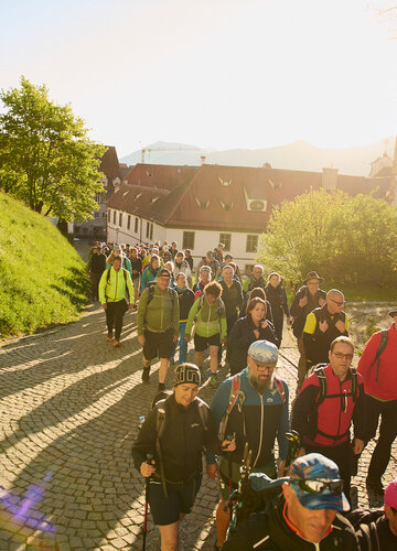 Das Bild zeigt eine Ansammlung von Menschen in Wanderbekleidung, die durch die Altstadt Füssens laufen. Hinter ihnen strahlt die Morgensonne.