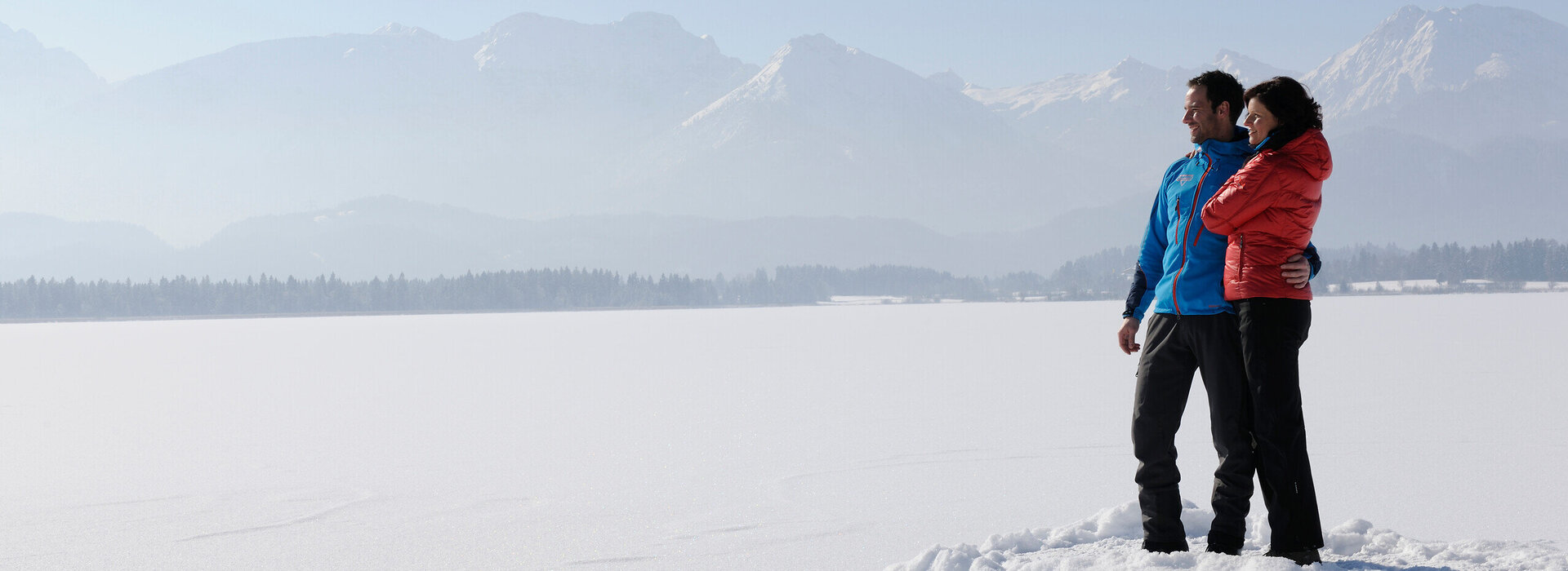 Das Bild zeigt einen Mann und eine Frau. Der Mann hat den Arm um die Frau gelegt. Sie stehen auf einem verschneiten Holzsteg. Der davorliegende See ist tief verschneit und nicht als See zu erkennen. Im Hintergrund erheben sich die schneebedeckten Berge. Die Landschaft ringsum ist tief verschneit. Der Himmel ist blau. Die Sonne scheint. 