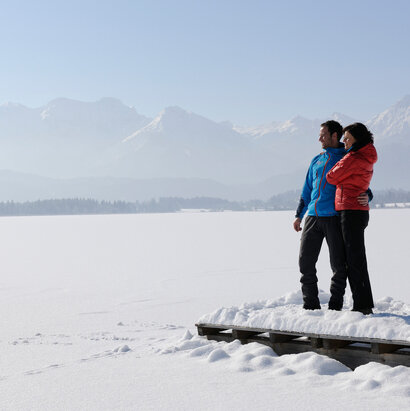 Das Bild zeigt einen Mann und eine Frau. Der Mann hat den Arm um die Frau gelegt. Sie stehen auf einem verschneiten Holzsteg. Der davorliegende See ist tief verschneit und nicht als See zu erkennen. Im Hintergrund erheben sich die schneebedeckten Berge. Die Landschaft ringsum ist tief verschneit. Der Himmel ist blau. Die Sonne scheint. 