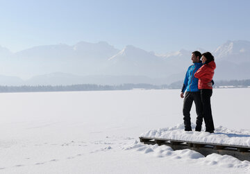 Das Bild zeigt einen Mann und eine Frau. Der Mann hat den Arm um die Frau gelegt. Sie stehen auf einem verschneiten Holzsteg. Der davorliegende See ist tief verschneit und nicht als See zu erkennen. Im Hintergrund erheben sich die schneebedeckten Berge. Die Landschaft ringsum ist tief verschneit. Der Himmel ist blau. Die Sonne scheint. 