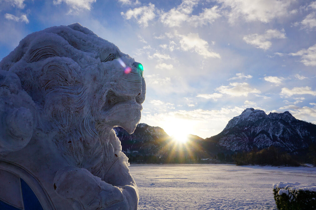 Eine Löwenskulptur aus Schnee steht im Vordergrund, während die Sonne hinter schneebedeckten Bergen aufgeht. Der Himmel ist teilweise bewölkt und zeigt eine Mischung aus Blau und Weiß. Die Winterlandschaft ist ruhig und friedlich.