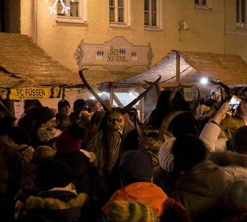 Das Foto zeigt ein große Menschenmenge, die in einer beleuchteten Gasse stehen. Mitten in der Menschenmenge sind zwei Klausen-Masken mit langen Hörnern und leuchtenden Augen zu sehen.   