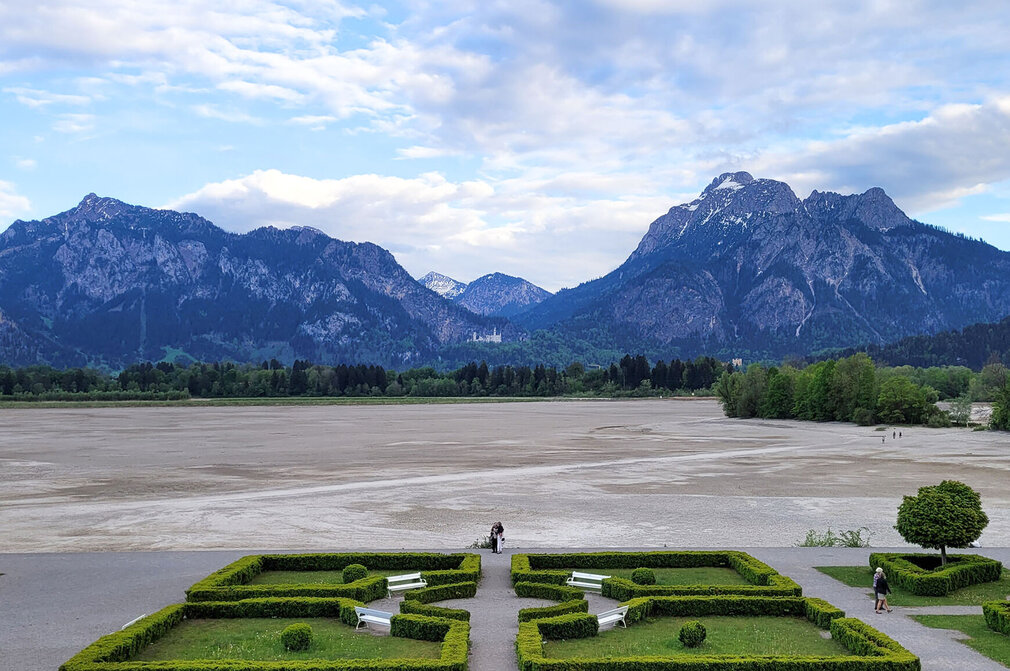 Ein weitläufiger Blick auf eine trockene Uferlandschaft mit Bergen im Hintergrund. Die Berge sind teils schneebedeckt und von Wolken umgeben. Im Vordergrund sind gepflegte, grüne Gartenbeete mit Sitzbänken zu sehen. Einige Personen sind auf dem trockenen Boden unterwegs.