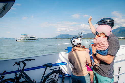 Das Bild zeigt eine friedliche Szene auf einem Boot. Eine Person mit einem pinken Helm hält ein Baby in den Armen. Das Baby trägt ebenfalls einen pinken Helm, was auf ein gemeinsames Interesse an Sicherheit hindeutet. Der Mann trägt außerdem einen pinken Helm, was darauf hindeuten könnte, dass er der Vater des Babys ist.  Das Boot, auf dem sie sich befinden, ist weiß und blau und liegt in einem Hafen. Der Hafen ist von Bergen umgeben, was der Szene einen Hauch von Natur verleiht. In der Ferne ist ein großes Boot zu sehen, was darauf hindeuten könnte, dass sie sich in einem beliebten Wassersportgebiet befinden.  Der Mann und das Baby sind nicht allein auf dem Boot. Auch ein Fahrrad ist vorhanden, was darauf hindeuten könnte, dass sie eine gemütliche Fahrt oder Exkursion unternehmen. Das Fahrrad steht auf dem Boot bereit, um verwendet zu werden, sobald sie ihr Ziel erreicht haben.  Insgesamt zeichnet das Bild das Bild einer Familie, die einen Tag auf dem Wasser genießt, mit Sicherheit an erster Stelle.