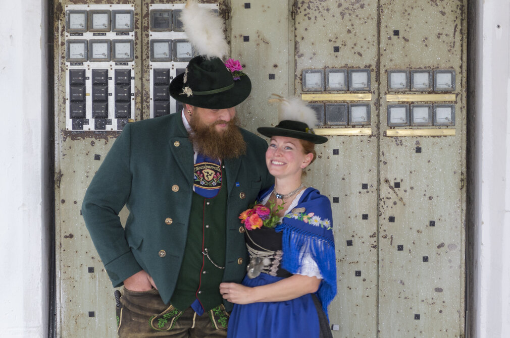 Ein Mann und eine Frau in traditioneller Tracht lächeln sich an. Der Mann trägt einen grünen Janker mit einem Hut, der eine Feder hat. Die Frau hat ein blaues Kleid mit bunten Blumen und einer Umhängetuch. Im Hintergrund sind alte Türen sichtbar.