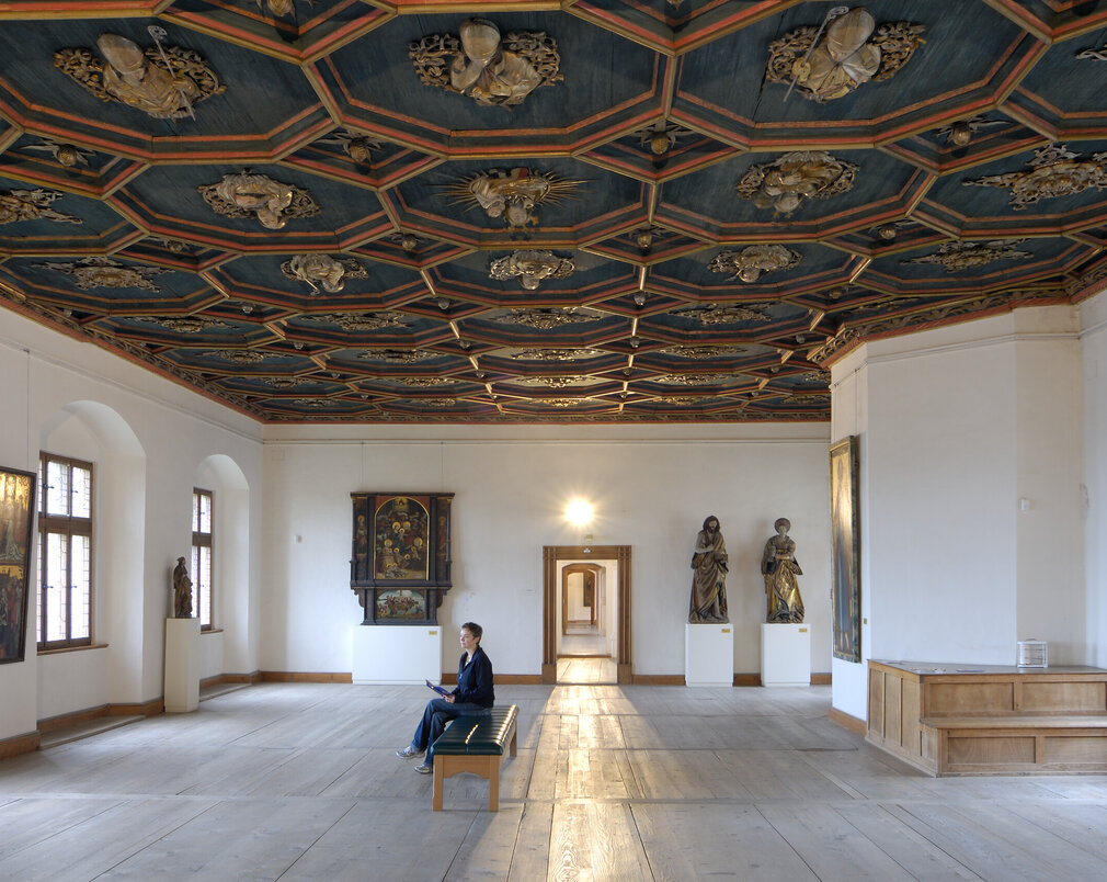 Man blickt in einen Saal mit weißen Wänden, einem historischen Holzboden und einer reich verzierten, dunkelblauen Holzdecke. Das Muster der Decke ist wabenförmig. Die Kanten der Waben sind erhaben und mit roten Umrandungen versehen. Innerhalb jeder Wabe ist eine, in Gold gearbeitete, kirchliche Person zu sehen. Man sieht Heiligenfiguren, aber auch Bischöfe und Päpste. An den Wänden hängen historische Gemälde mit unterschiedlichen Themen. Der Raum ist, bis auf eine moderne mit schwarzem Leder bezogene Bank in der Mitte, nicht möbliert. Auf der Bank sitzt eine Frau und blickt auf die Gemälde. 