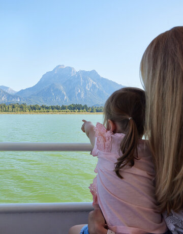 Das Bild fängt eine friedliche Szene von zwei Menschen ein, die mit Kajaks auf einem See unterwegs sind. Die Kajakfahrer tragen Schwimmwesten und paddeln in einem roten Kajak. Der See ist von majestätischen Bergen umgeben, was der Szene ein Abenteuergefühl verleiht. Der Himmel darüber ist ein klarer Blau, was auf einen schönen Tag für Outdoor-Aktivitäten hindeutet. Die Gesamtatmosphäre des Bildes ist eine von Ruhe und Naturgenuss.