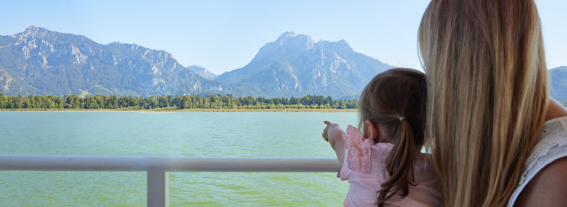 Das Bild fängt eine friedliche Szene von zwei Menschen ein, die mit Kajaks auf einem See unterwegs sind. Die Kajakfahrer tragen Schwimmwesten und paddeln in einem roten Kajak. Der See ist von majestätischen Bergen umgeben, was der Szene ein Abenteuergefühl verleiht. Der Himmel darüber ist ein klarer Blau, was auf einen schönen Tag für Outdoor-Aktivitäten hindeutet. Die Gesamtatmosphäre des Bildes ist eine von Ruhe und Naturgenuss.