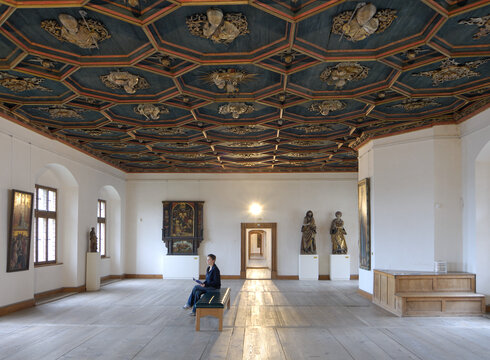 Der Raum hat eine hohe Decke mit einem dunkelblauen Deckenfresko und Holzdielen. Eine Person sitzt auf einer Bank, während Statuen und ein Gemälde im Raum ausgestellt sind.