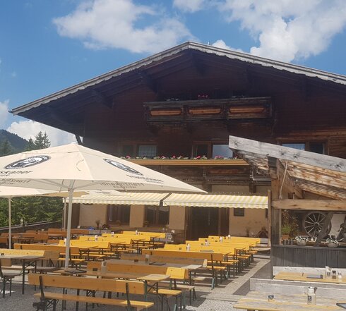 DAs Bild zeigt ein Haus mit großen Holzbalkonen, auf denen bunte Blumen blühen. Auf der davorliegenden Terrasse stehen aufgespannte, weiße Schrime. Im Hintergurn sind Nadelbäume zusehen.