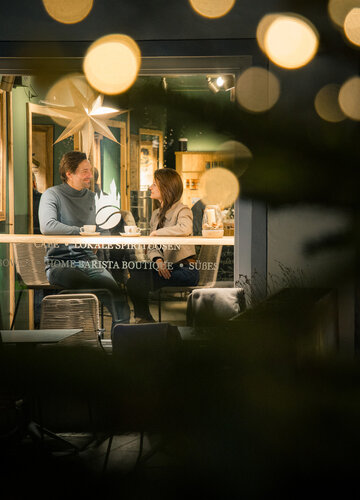 Blick durch ein Cafe-Fenster auf ein Paar an einem Tisch mit Tassen, vor unscharfen, funkelnden Lichtern und einer großen Stern-Dekoration.