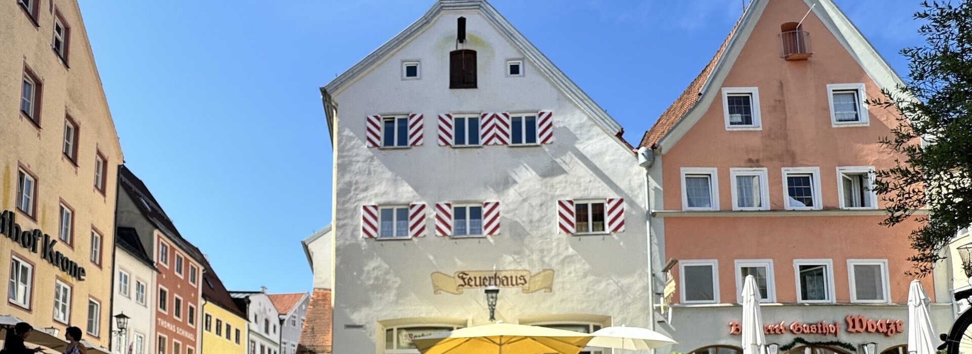 Dieses Bild zeigt einen belebten Platz in der historischen Altstadt mit farbenfrohen Fachwerkhäusern. Im Vordergrund befindet sich ein markantes Gebäude mit einem Turm und einem Zifferblatt, das von Menschen, die draußen sitzen und essen, umgeben ist.