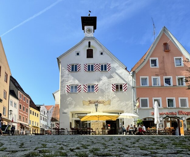 Dieses Bild zeigt einen belebten Platz in der historischen Altstadt mit farbenfrohen Fachwerkhäusern. Im Vordergrund befindet sich ein markantes Gebäude mit einem Turm und einem Zifferblatt, das von Menschen, die draußen sitzen und essen, umgeben ist.