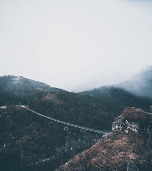 Das Bild zeigt eine atemberaubende Landschaft mit Gebirgen. Die Szene wird von einer windenden Straße dominiert, die sich wie eine Schlange durch das Gelände schlängelt und in der Ferne verschwindet. Die Straße, ein Faden aus grauem Asphalt, ist von saftig grünen Bäumen und Sträuchern flankiert, deren leuchtende Farben schön im Kontrast zum Grau der Straße stehen. Der Himmel darüber ist ein helles Blau, durchzogen von zarten Wolken, die der Szene Tiefe und Weite verleihen. Die Perspektive des Bildes ist eine hohe Aufsicht, die auf die Straße und die umgebende Landschaft hinabschaut. Diese Perspektive erlaubt eine umfassende Sicht auf das Gebiet und zeigt den windenden Pfad der Straße und die dichte Vegetation, die sie säumt. Die Gesamtkomposition des Bildes erzeugt ein Gefühl der Ruhe und Ehrfurcht und fängt die natürliche Schönheit der Gebirgslage ein.